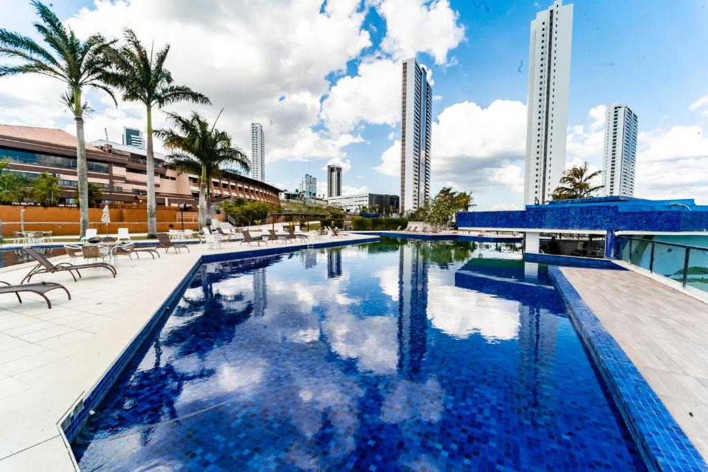 Piscina grande e azul com espreguicadeiras ao lado, palmeiras e prédios vistos ao fundo.