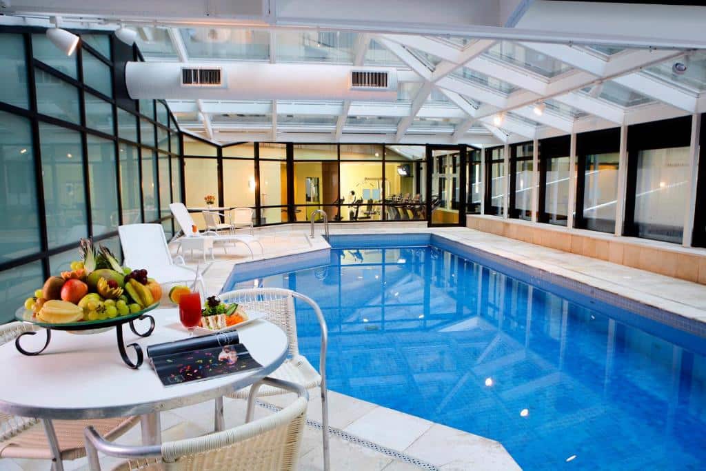 Área da piscina interna coberta do hotel Bourbon, é separada por um vidro com a academia do hotel. Há uma mesa com frutas e várias espreguiçadeiras ao redor da piscina.