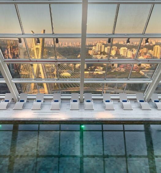 Piscina coberta do Hilton São Paulo Morumbi. O teto é de vidro, em formato de triângulo, sendo possível ver desde sua base toda a vista para a cidade de São Paulo. Há várias espreguiçadeiras e uma escada de acesso para a piscina, que possui luzes dentro.