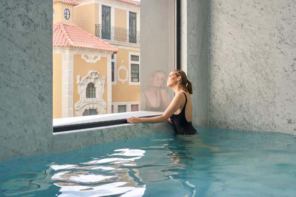 Piscina coberta do PortoBay Flores com uma mulher loira dentro da piscina olhando a janela com vista para a cidade.