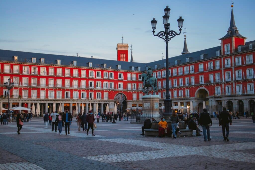 Pessoas passeando pela Plaza Mayor em Madri. Uma construção vermelha repleta de janelas cerca a praça de todos os lados, e uma estátua de um homem montado em seu cavalo está no centro do local. Algumas pessoas estão sentadas na base de um poste de luz enquanto conversam. - Foto: Eduardo Rodriguez via Unsplash