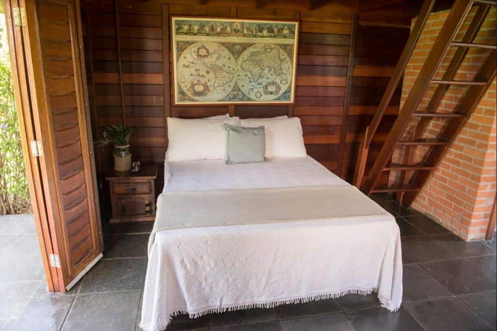 Chalé com paredes de madeira, cama de casal com lençois brancos, quadro decorativo acima da cama, porta e escada de madeira nos lados da cama. Imagem para ilustrar o post hotéis e pousadas em Itu.
