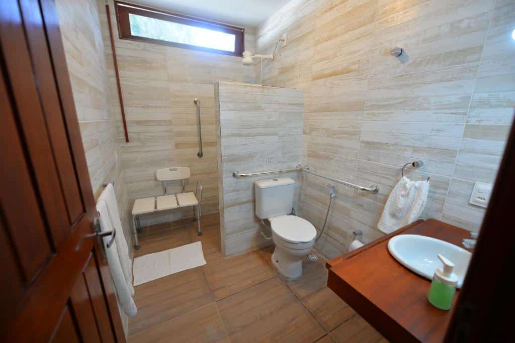 Banheiro da Pousada do Gunga com uma pia, um vaso com duas barras de apoio ao redor e área do chuveiro com uma barra de apoio e uma cadeira de banho.