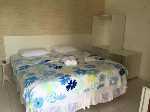 Quarto da Pousada Villa Joia com uma cama de casal com toalhas em cima e um armário ao lado. Foto para ilustrar post sobre pousadas em Barra de de São Miguel.