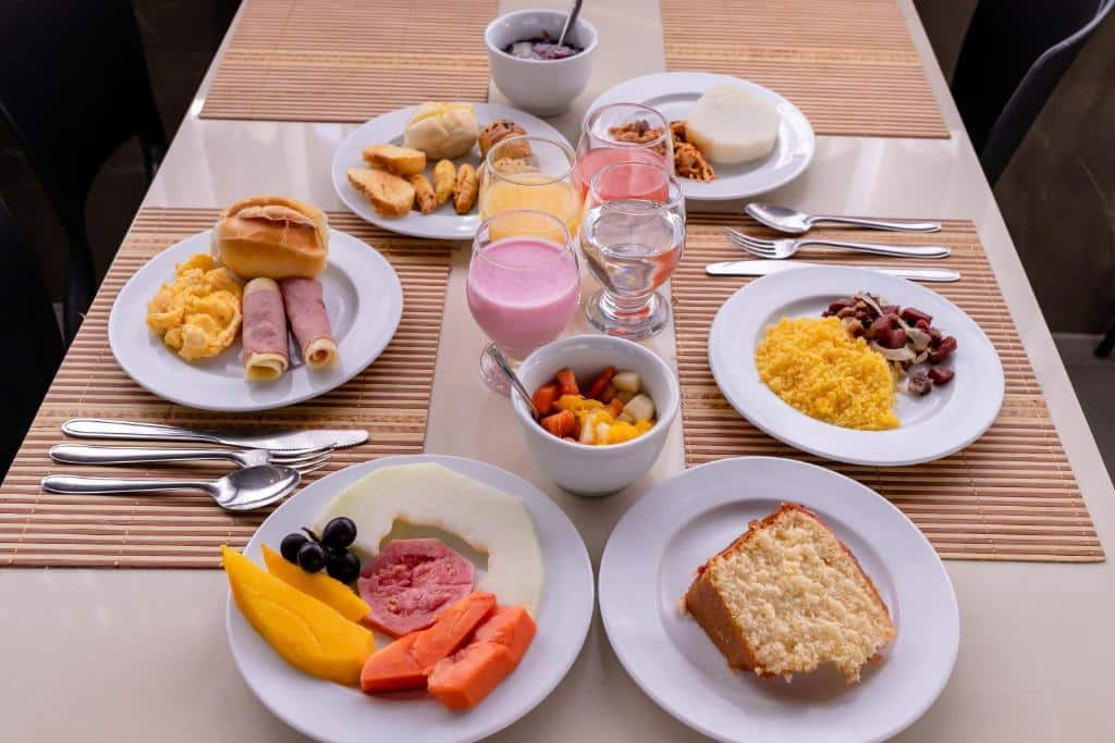 Mesa com café da manhã com pratos de frutas, bolo, pães, ovos, cuscuz, salada de frutas, copos com suco, iogurte e água.