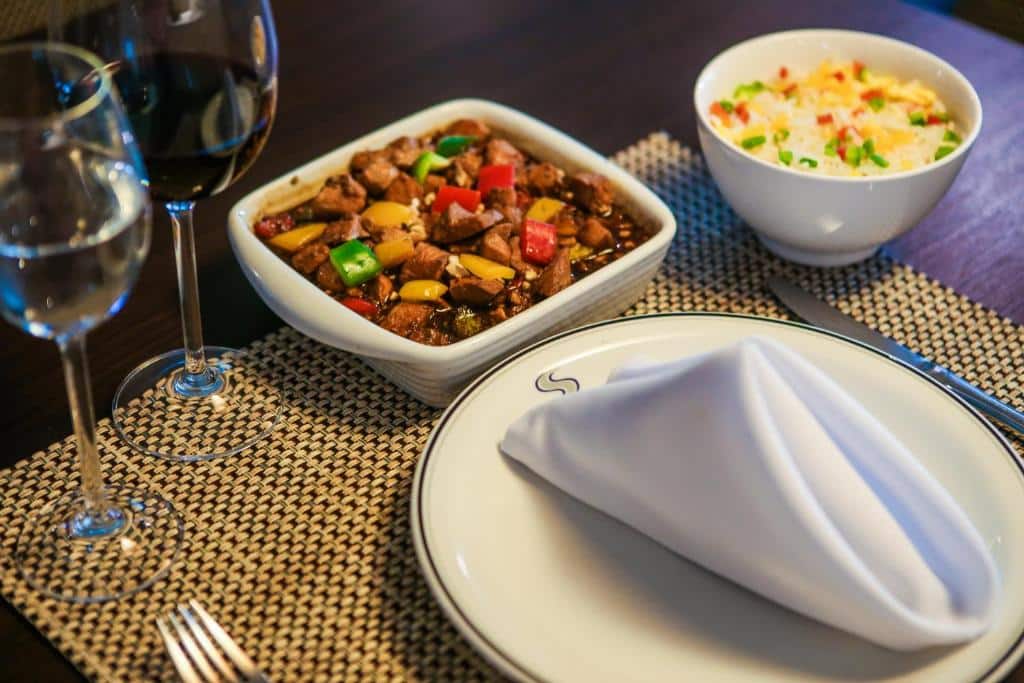 Prato branco com guardanapo de tecido branco em cima, taças de vinho e água e talheres ao lado, pratos com carnes e arroz.