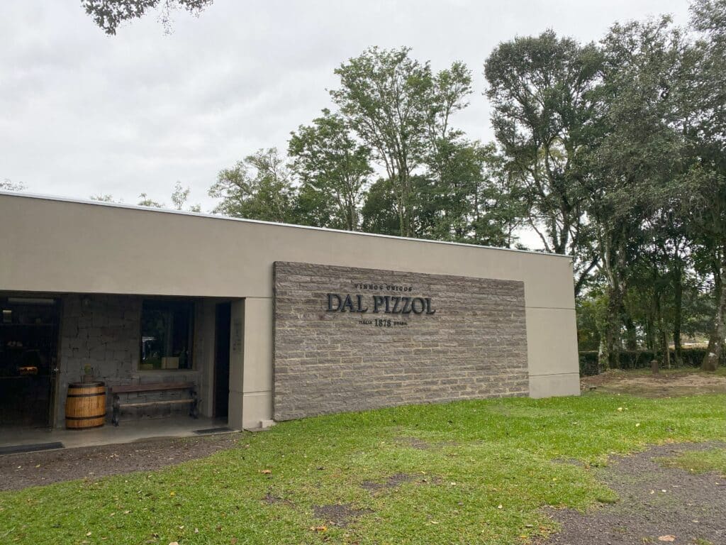 Prédio da vinícola Dal Pizzol, uma das vinícolas no Vale dos Vinhedos, onde fica um museu sobre o vinho brasileiro, com letreiro do nome da marca em parede de pedra, além de barril de madeira ao lado de um banco em frente à porta de entrada