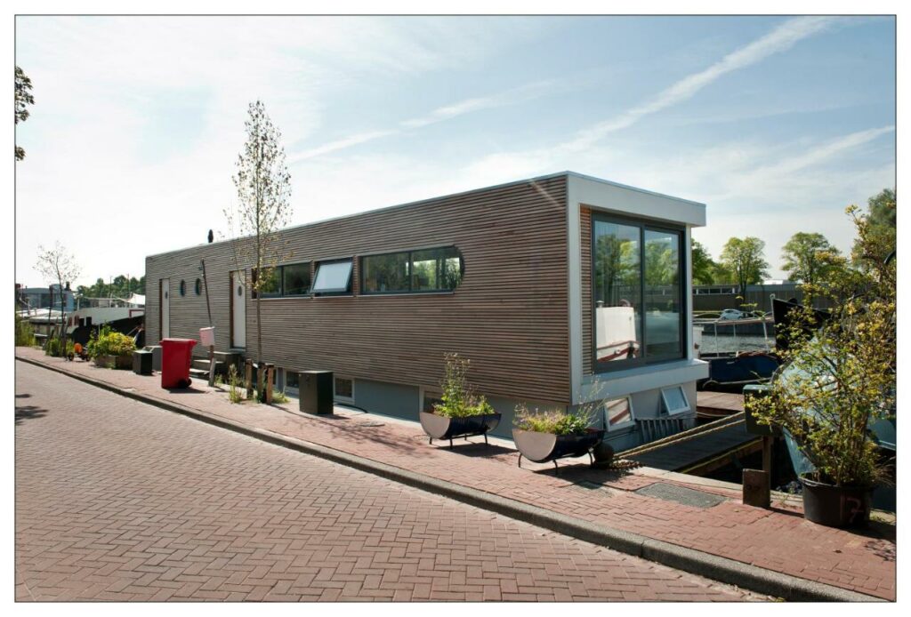 Um dos airbnb em Amsterdam, sendo o local uma casa flutuando no formato de um retangulo grande, localizado na beira de um rio, com janelas de vidro, caminhos em volta de madeira e um vasos com flores e plantas na frente