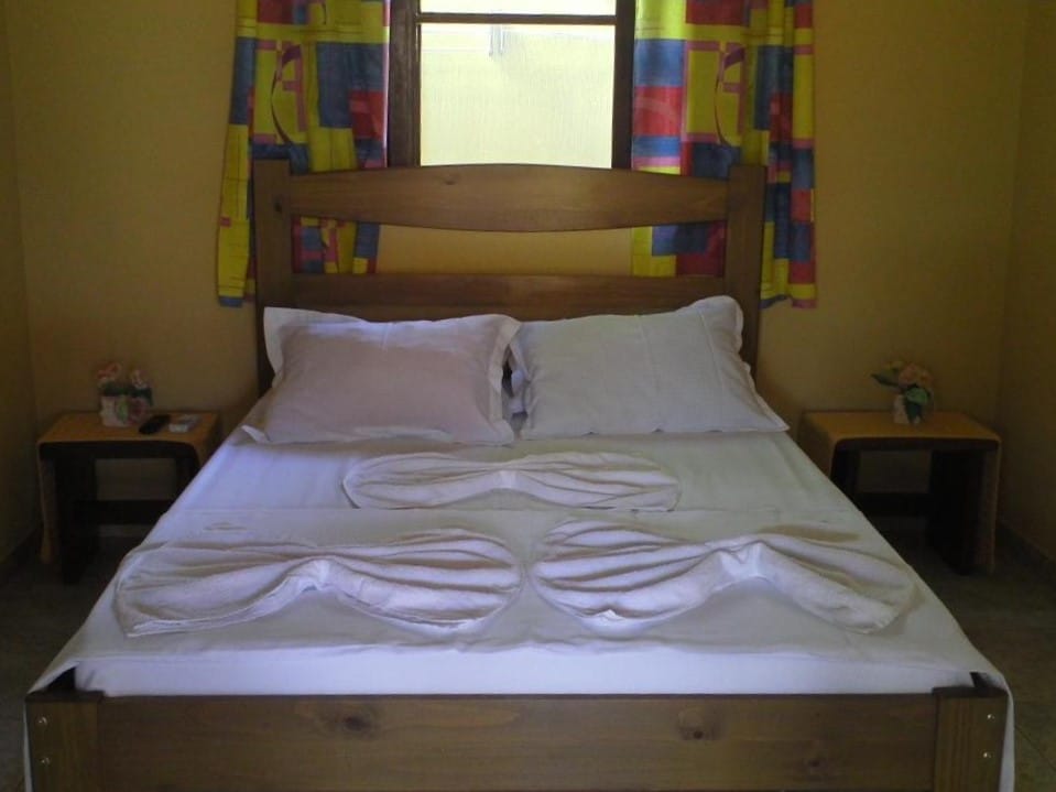 Uma cama de casal no Makitub. Atrás da cama uma janela com cortina. Uma prateleira de apoio em cada lado da cama.