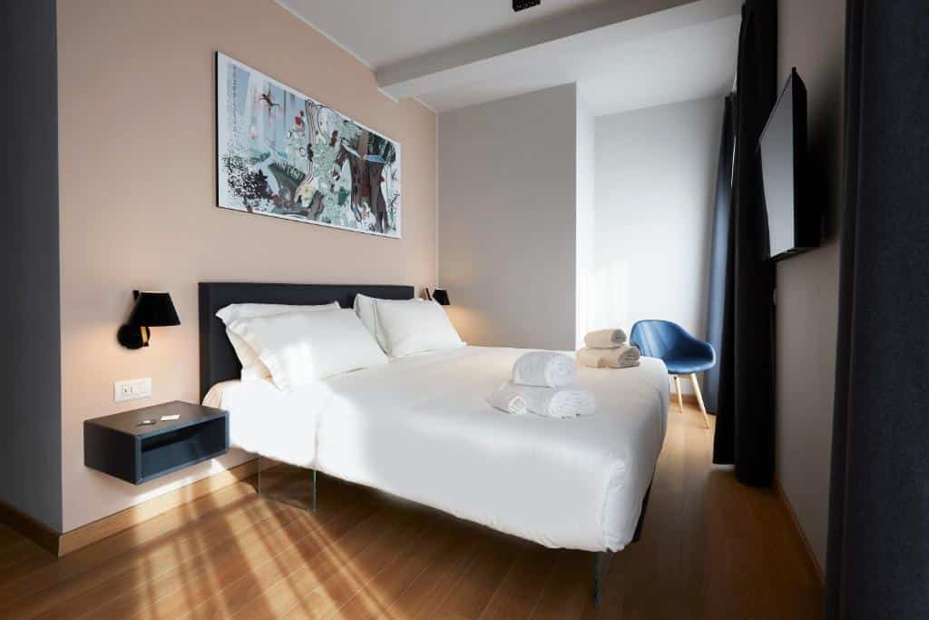 Quarto do 21 House of Stories Città Studi com uma cama de casal, uma televisão, chão que imita madeira, uma cadeira próxima da janela com cortinas, e uma mesinha de cabeceira com uma luminária, para representar os melhores hotéis em Milão