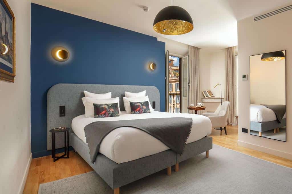 quarto do 9Hotel Cesari, um dos hotéis perto da Fontana di Trevi em Roma, com cama de casal, mesinha e luminária de ambos os lados, lustre acima redondo, parede e detalhes em azul acinzentado, poltrona branca com mesinha e janela comprida com vista