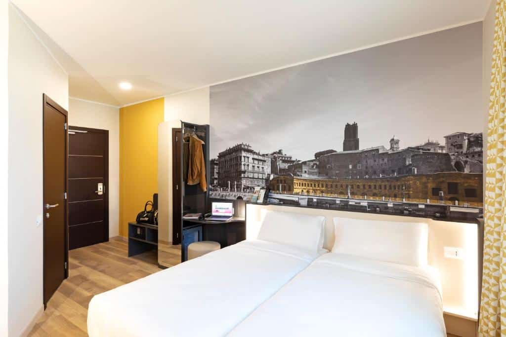 quarto do B&B Hotel Roma Fiumicino Aeroporto Fiera 1, um dos hotís perto do aeroporto em Roma, com duas camas de solteira unidas, com um quadro da cidade atrás, guarda-roupa, mesinha de cabeceira e vários luminárias