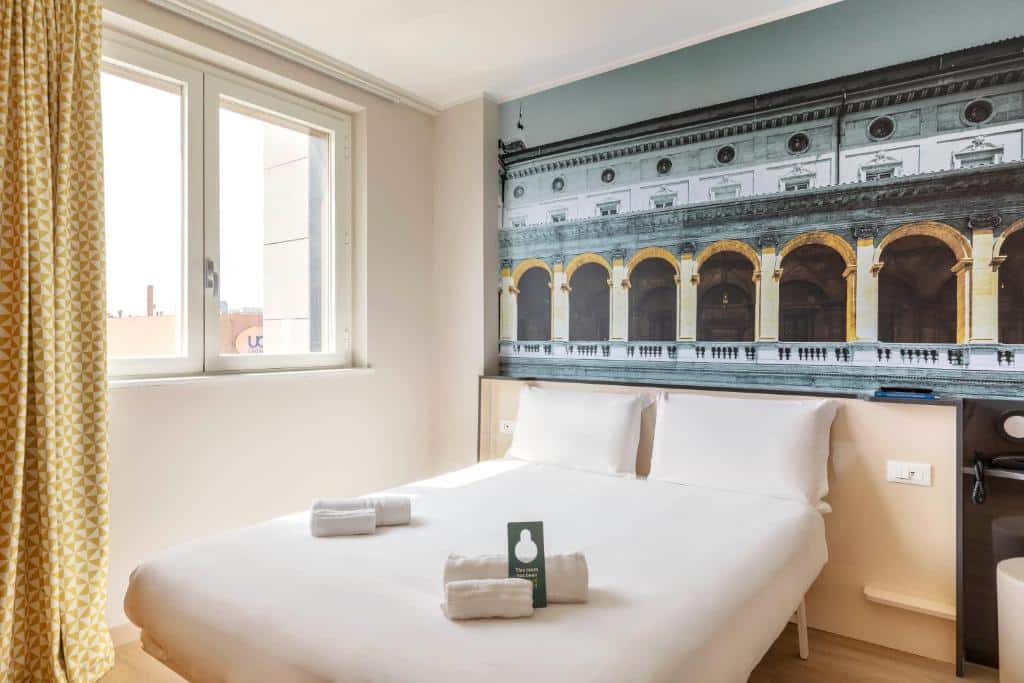 quarto do B&B Hotel Roma Fiumicino Aeroporto Fiera 2, um dos hotéis perto do aeroporto de Roma, com cama de casal com toalhas acima, quadro do Coliseu acima e janela quadrada do outro lado