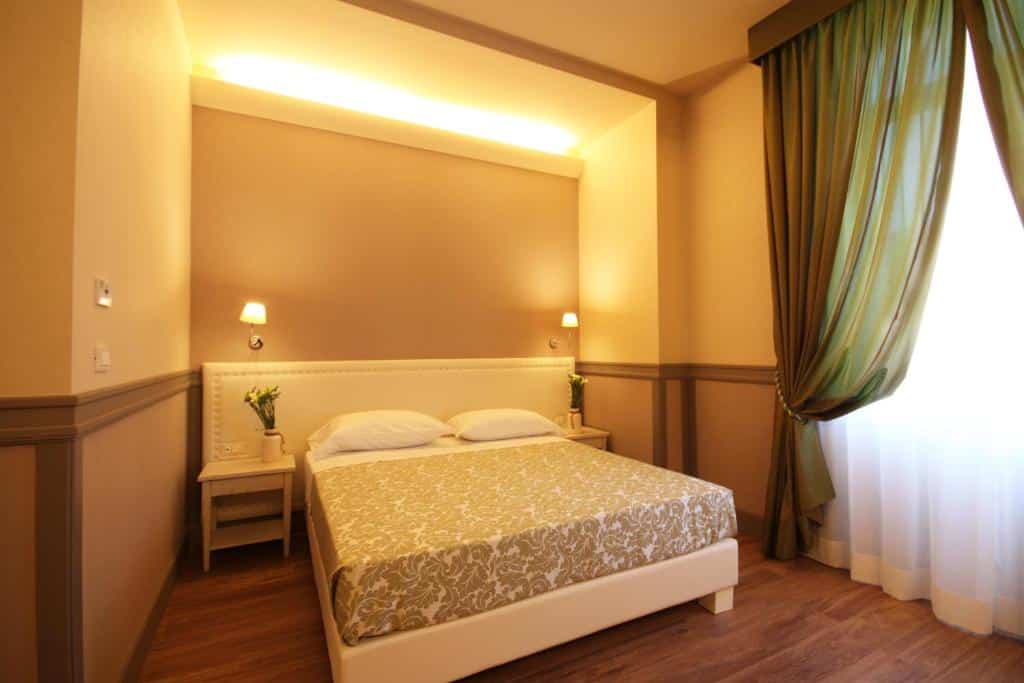 quarto do Basilica Hotel, um dos hotéis bem localizados em Roma, com cama de casal, mesinha e luminária de ambos os lados, roupa de cama florida com janela ao lado com cortinas leves