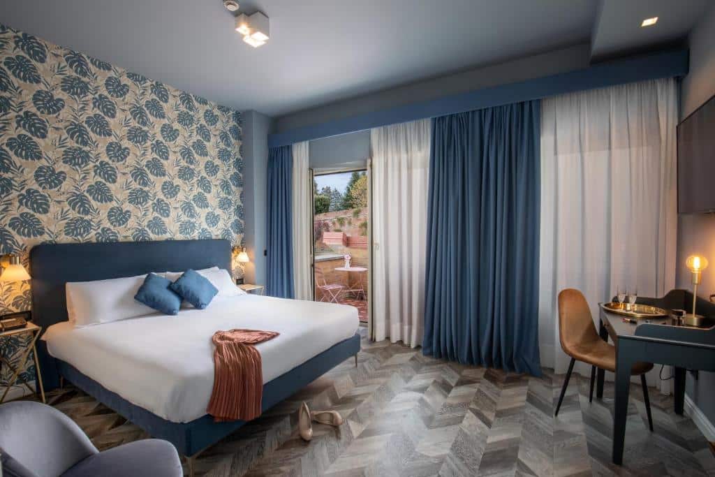 quarto do Bloom Hotel Rome, um dos hotéis perto do Vaticano, com cama de casal, mesinha e luminária de ambos os lados, poltrona e mesa à frente com tv, além de poltrona e janela grande com cortinas e varanda