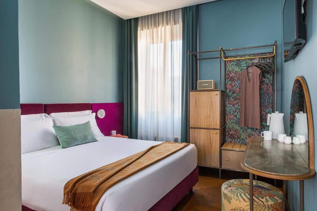 quarto do Condominio Monti Boutique Hotel, um dos hotéis perto do Coliseu em Roma, com cama de casal, mesinha e luminária de ambos os lados, com detalhes em verde suave, arroxeados e em madeira, com guarda-roupa aberto, mesa e cadeiras, há uma janela com cortinas leves