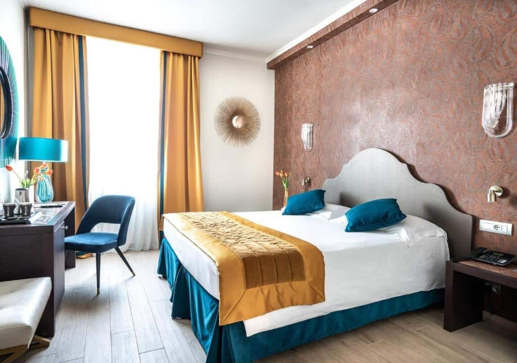 quarto do FH55 Grand Hotel Palatino, um dos hotéis para brasileiros em Roma, com cama de casal, mesinha e luminária de ambos os lados, quadro em forma de sol na parede, poltrona, mesa e tv à frente, há uma janela grande com cortinas, os detalhes são em azul e dourado