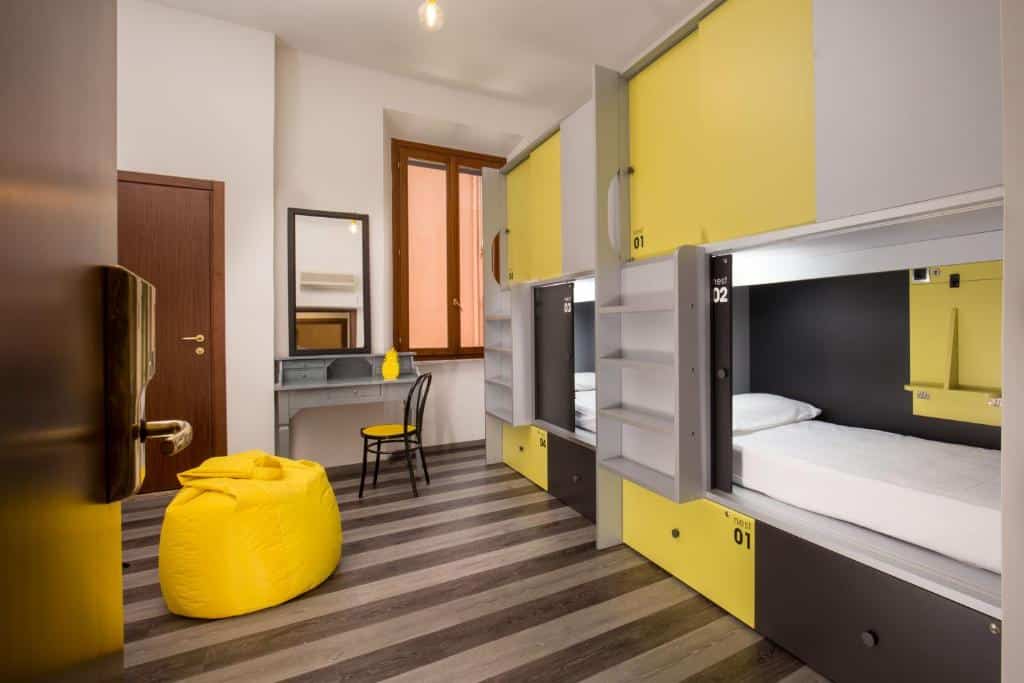 quarto do Free Hostels Roma, com mesa, cadeira e espelho, puff amarelo e duas beliches também amarelas, com escadinha e cofre com chave, há uma janela de madeira