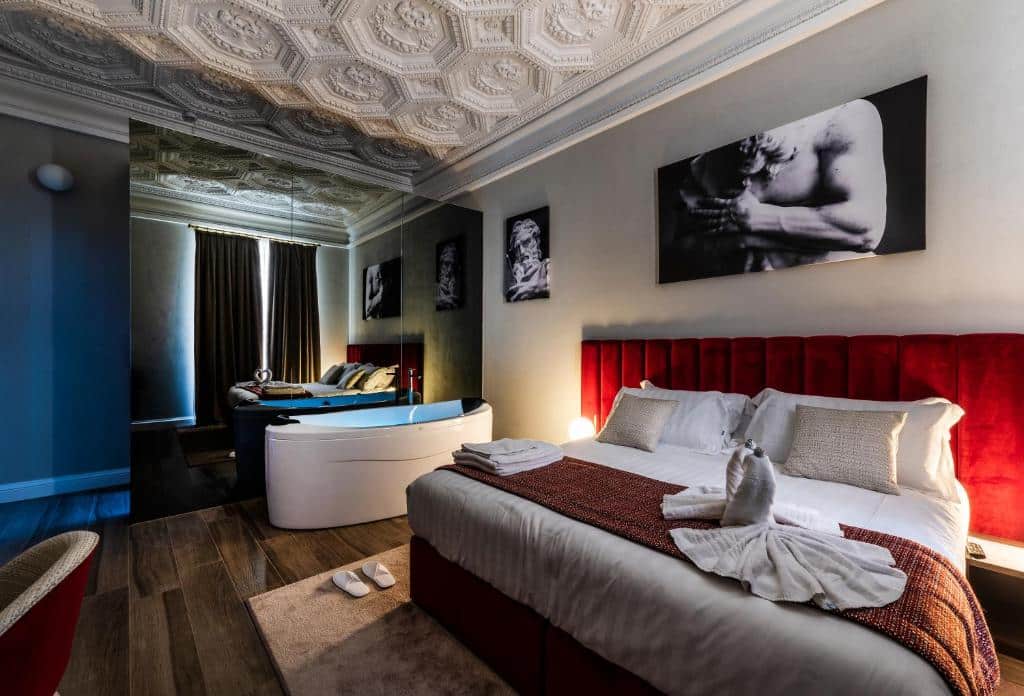 quarto do Growel Exclusive Suites San Pietro, um dos hotéis românticos em Roma, com cama de casal enome, banheira de hidro oval com um grande espelho, há quadros na parede e o teto tem flores desenhadas em gesso, há ainda mesinha e luminária de ambos os lados e potrona