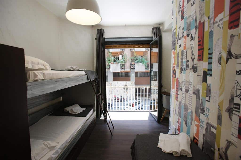 quarto do Hostel Trastevere, um dos hostels em Roma, com uma beliche simples e mesa à frente com papel de parede colorido e varanda vista de uma porta grande de madeira