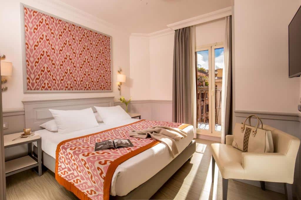 quarto do Hotel Della Conciliazione, um dos hotéis para brasileiros em Roma, com cama de casal, mesinha e luminária de ambos os lados, mesa e cadeira à frente, com colcha e papel de parede combinando em detalhes vermelhos, há uma janela com vista e cortinas