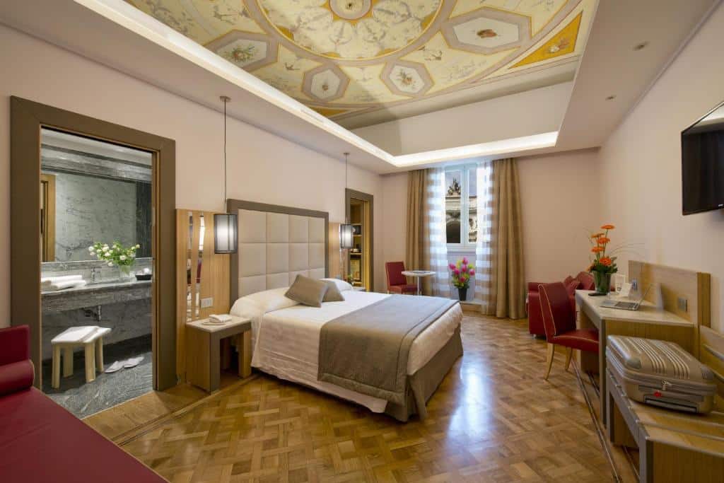 quarto do Hotel Giolli Nazionale, um dos hotéis para brasileiros em Roma, em uma ambiente com cama de casal, mesinha e luminária de ambos os lados, mesa e poltrona à frente, janela grande com cortinas, e entrada para o banheiro e teto trabalhado com pinturas amarelas