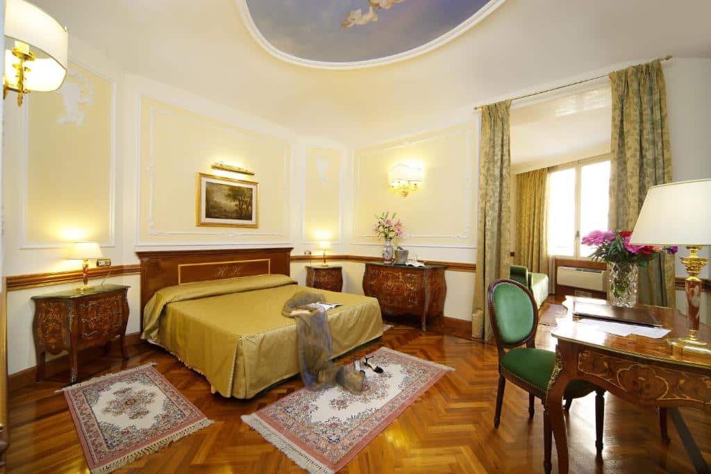 quarto do Hotel Hiberia, um dos hotéis bem localizados em Roma, com cama de casal, mesinha e luminária de ambos os lados, mesa e poltrona à frente, com tapetes no chão de madeira que combina com todos os móveis, também em madeira, há uma janela grande com cortinas e decoração clássica