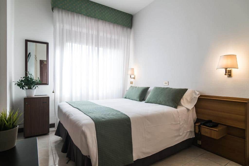 quarto do Hotel Pacific um dos hotéis perto do Vaticano, com cama de casal, com travesseiros verdes, combinando com detalhes das cortinas, há mesinha e luminária de ambos os lados da mesa, e a janela é grande, há um aparador e espelho na parede