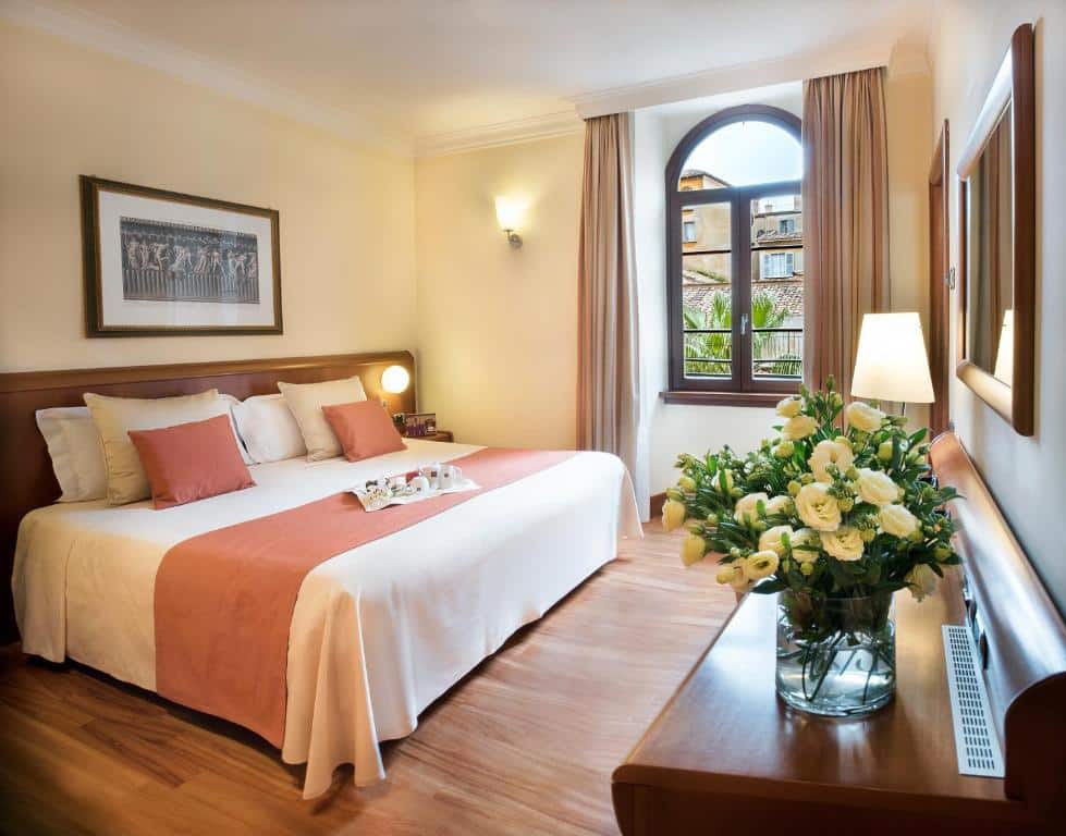 quarto do Hotel Ponte Sisto, um dos hotéis para brasileiros em Roma, com cama de casal grande, mesinha e luminária de ambos os lados, janela de madeira com bordas arredondadas e cortinas, com mesa à frente com um vaso de flores