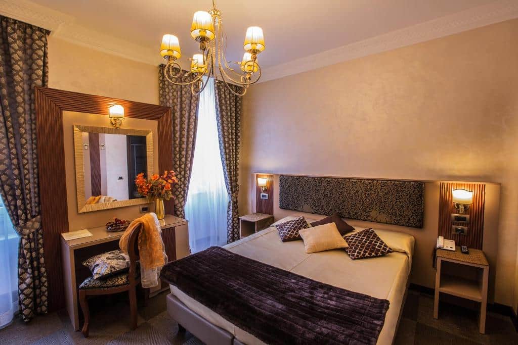 quarto do Hotel Romano, com cama de casal, mesinha e luminária de ambos os lados, penteadeira com poltrona e lustre de teto com decoração clássica