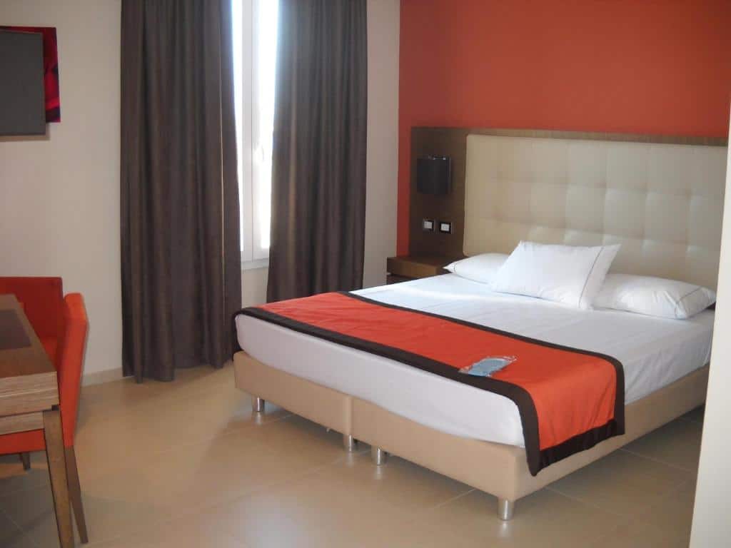 quarto do Hotel Tiber, um dos hotéis perto do aeroporto em Roma, com cama de casal, mesinha e luminária em ambos os lados, mesa, cadeira e tv à frente e uma janela comprida do outro lado, os detalhes são em laranja