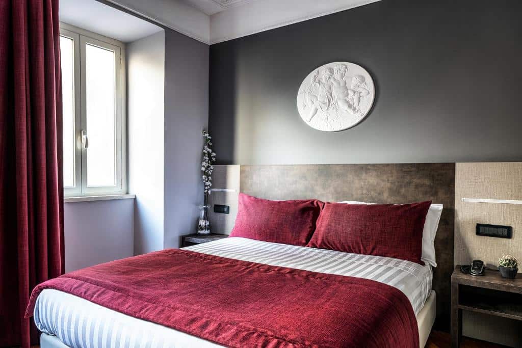 quarto do Hotel Varese, um dos hotéis bem localizados em Roma, com cama de casal com detalhes vermelhos, mesinhas e luminária de ambos os lados, quadro redondo com esculturas trabalhadas em relevo com janela grande e cortinas