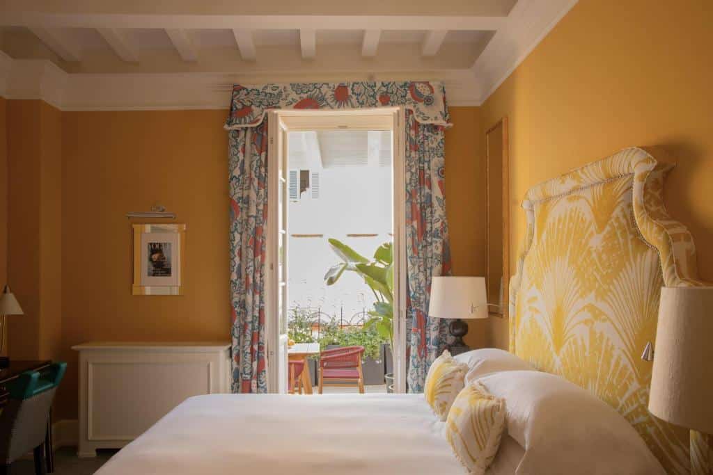 quarto do Maalot Roma - Small Luxury Hotels of the World, um dos hotéis perto da Fontana di Trevi em Roma, com cama de casal grande, mesinha e luminária de ambos os lados, com mesa e cadeiras na varanda, há uma janela grande com cortinas florida e paredes amarelas