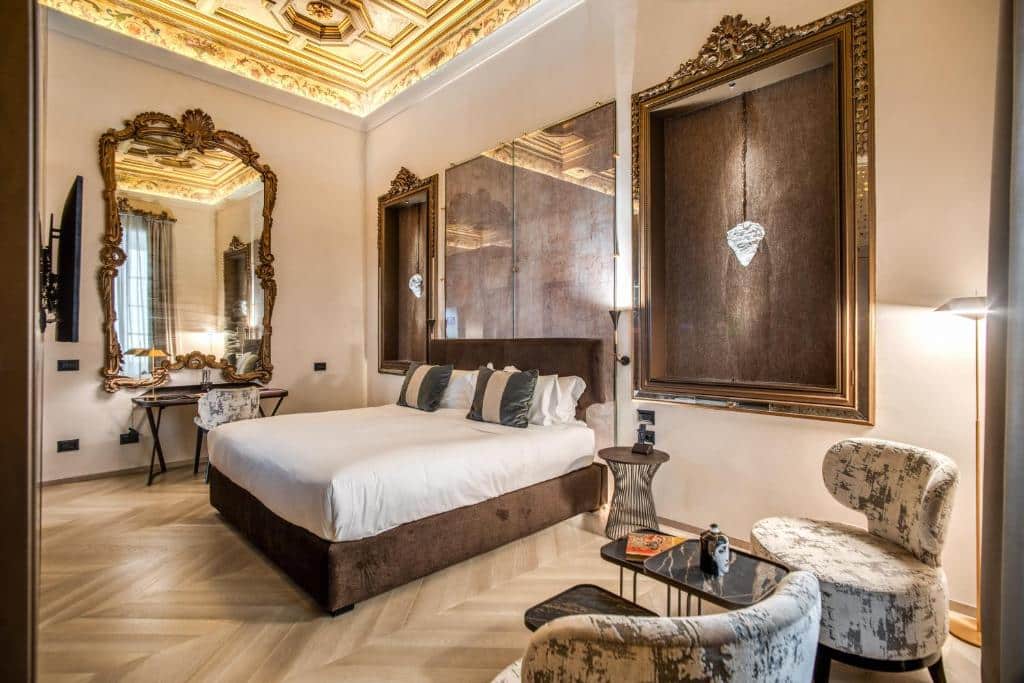 quarto do Martius Private Suites Hotel, um dos hotéis românticos em Roma, com cama de casal, luminárias de ambos os lados, há ainda duas poltronas com estampa de flores e tudo em decoração clássica, do outro lado, um espelho enorme com moldura muito trabalhada e tv à frente