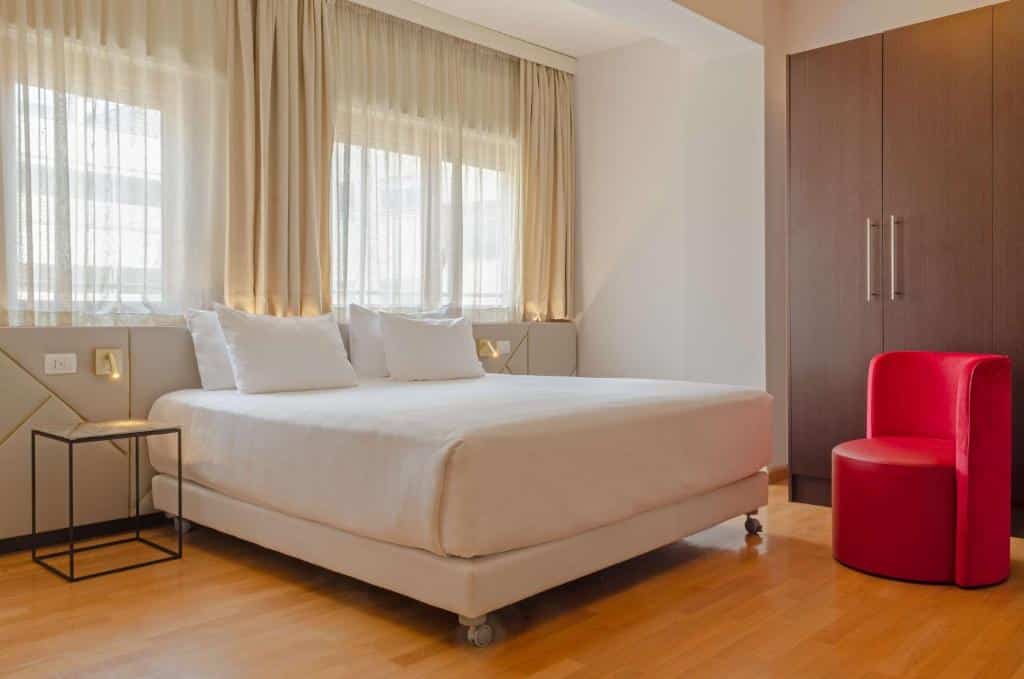 quarto do NH Collection Roma Giustiniano, um dos hotéis perto do Vaticano, com cama de casal, mesinha e luminária de ambos os lados, janela com cortinas claras acima da cama, com guarda-roupa de madeira a poltrona vermelha
