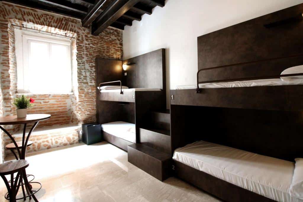 quarto do New Generation Hostel Rome Center, com duas beliches pretas, camas simples uma escada no centro, há uma mesa com cadeira e janela em uma parede que imita tijolinho à mostra