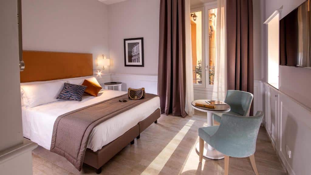 quarto do Princeps Boutique Hotel, um dos hotéis bem localizados em Roma, com cama de casal, mesinha e luminária de ambos os lados, janela grande com cortinas e poltronas com uma mesa pequena e tv à frente
