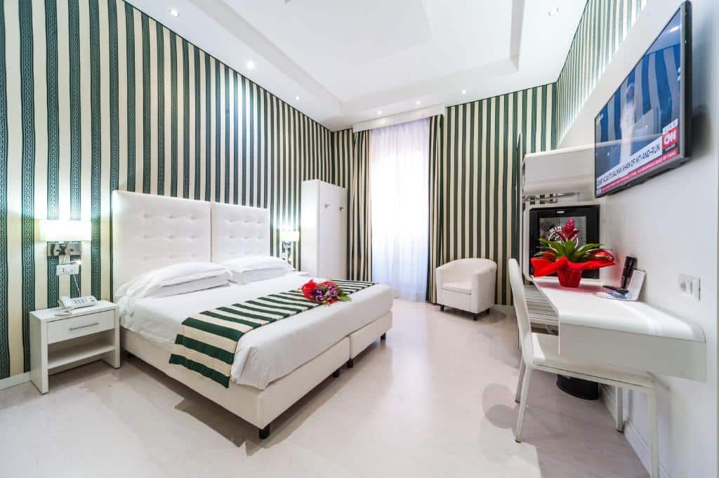 quarto do Relais Trevi 95 Boutique Hotel, um dos hotéis perto da Fontana di Trevi em Roma, com cama de casal grande, mesinha e luminária de ambos os lados, tv, bancada e poltrona à frente e poltrona ao lado, todos os móveis são brancos com bordas arredondadas, as paredes e aos detalhes da cama são listrados com verde e branco