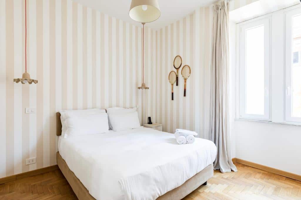 quarto do Salotto Monti, um dos hotéis bem localizados em Roma, com cama de casal com luminária de ambos os lados tudo em branco e detalhes muito pontuais dourados, janela grande com cortinas pesadas, com raquetes ornamentais na parede e papel de parede listrado