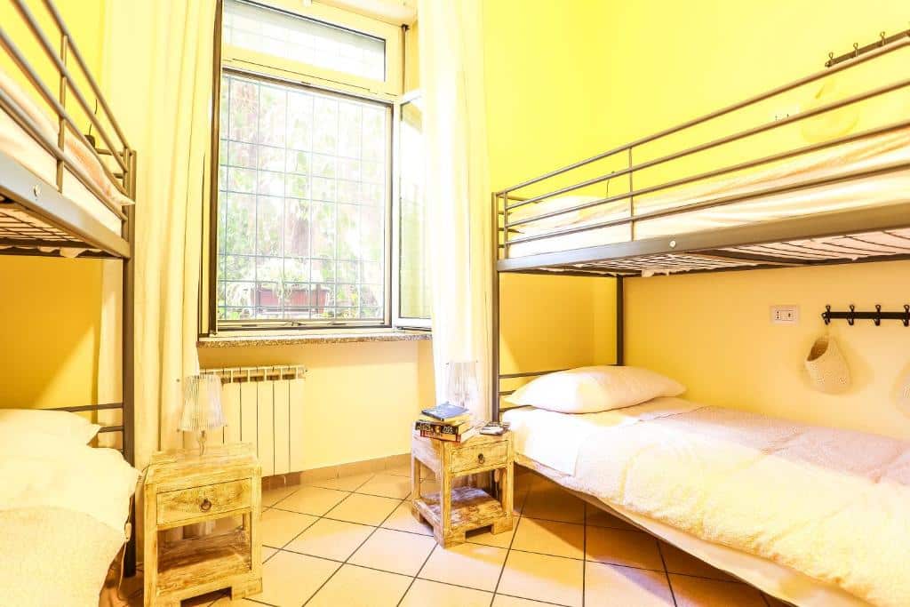 quarto do The Beehive, um dos hostels em Roma, com duas beliches simples, há um móvel ao lado com luminária e uma escadinha, janela com cortinas