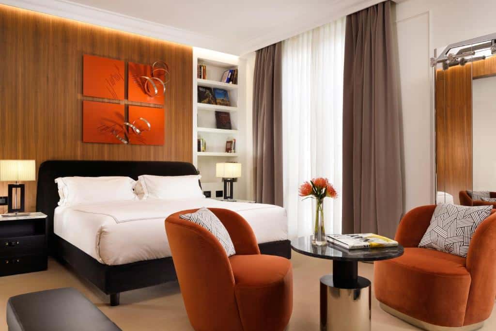 quarto do The First Dolce - Preferred Hotels & Resorts, um dos hotéis para brasileiros em Roma, com cama de casal, mesinha e luminária de ambos os lados, prateleiras com decoração, poltronas laranjas e mesinha de centro e janela bem grande com cortinas