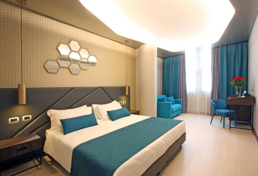quarto do The Hive Hotel, um dos hotéis bem localizados em Roma, com cama de casal bem grande, mesinha e luminária de ambos os lados, espelho em formato de colmeia acima, e sala de estar com sofá e poltrona do outro lado com janela grande e cortinas, a cor dos móveis é azul e há luzes no teto