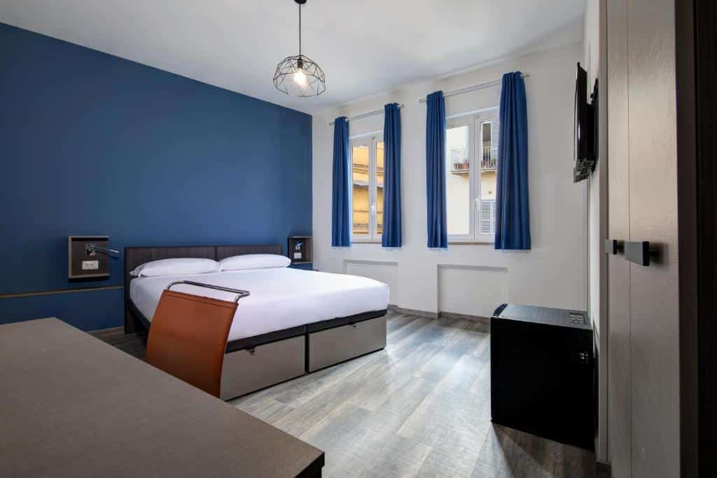 quarto do hostel do The RomeHello com cama de casal com mesinha e luminária de ambos os lados, duas janelas com cortinas azuis, combinando com a parede da mesma cor, há uma luminária no teto, mesa com cadeira e baú