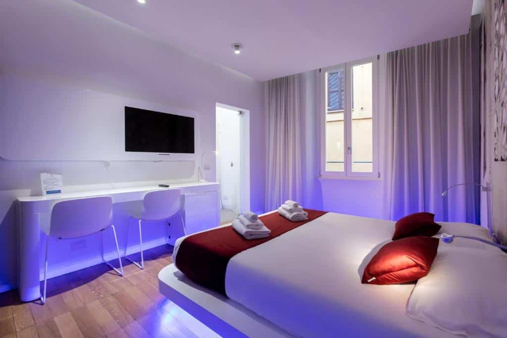quarto do Trevi Elite Rome, um dos hotéis perto da Fontana di Trevi em Roma, com cama de casal e luzes abaixo neon azuis, com tv, poltrona e guarda-roupa aberto de design, tudo em branco com janela e cortinas claras