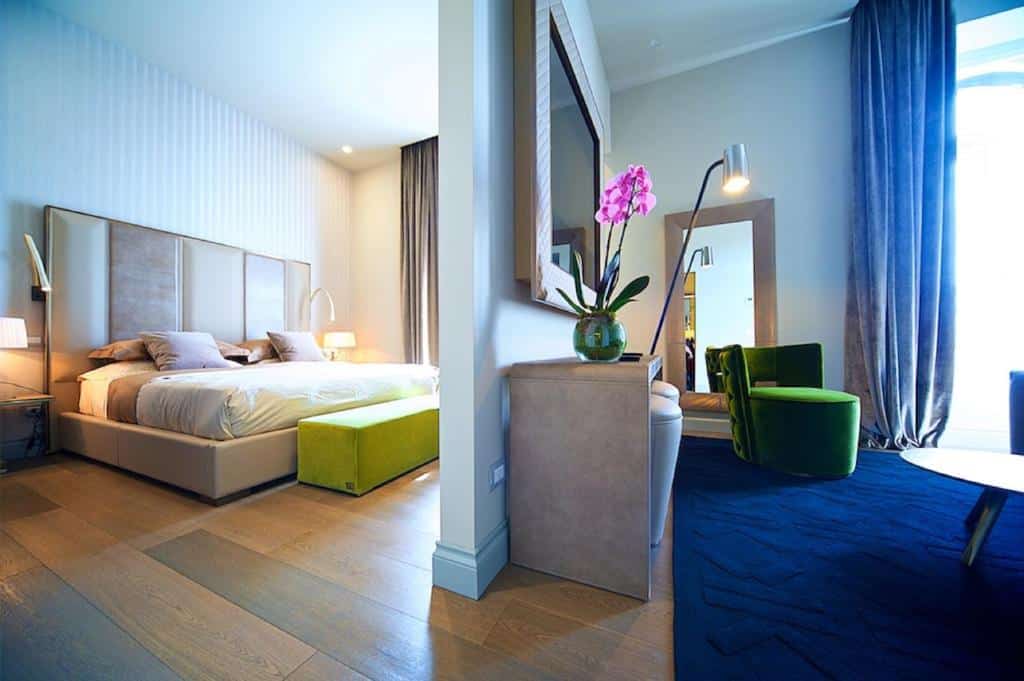 quarto do Vittoriano Luxury Suites, um dos hotéis românticos em Roma, com cama king size, mesinha e luminária de ambos os lados e uma área de estar com tv, sofá e poltronas em tons de azul e verde