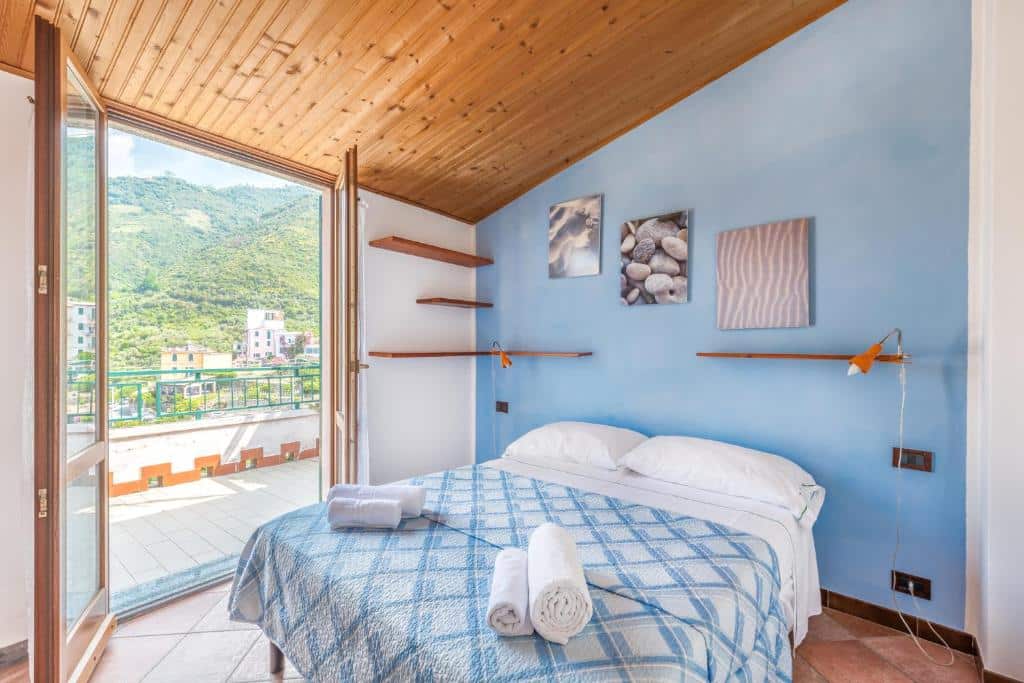 Quarto com uma cama de casal com detalhes azuis e brancos, paredes nas mesmas cores, teto de madeira, três quadros na parede e uma porta grande que dá acesso a varanda.