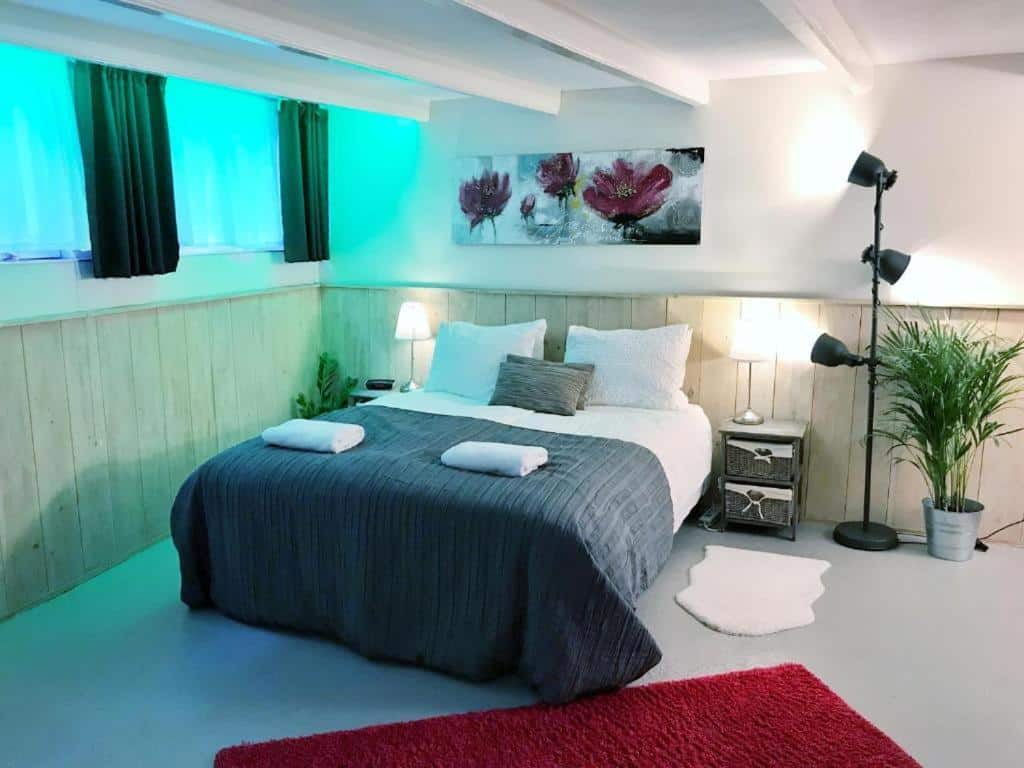 Quarto do Amsterdam Home, com uma cama de casal com travesseiros e toalhas em cima, um tapete vermelho a frente e, no lado da cama, tem uma escrivaninha, uma luminária vertical e um vaso de flor. Representa airbnb em Amsterdam