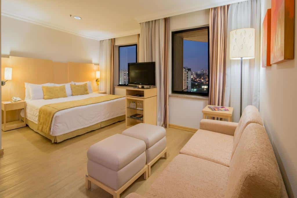 Imagem do quarto do Blue Tree Premium Morumbi, para o post sobre hotéis com piscina aquecida em São Paulo. A suíte é ampla e comporta uma cama box de casal, uma estante com TV, dois puffs em formato quadrado e um sofá. Há duas janelas no quarto, do lado da cama.