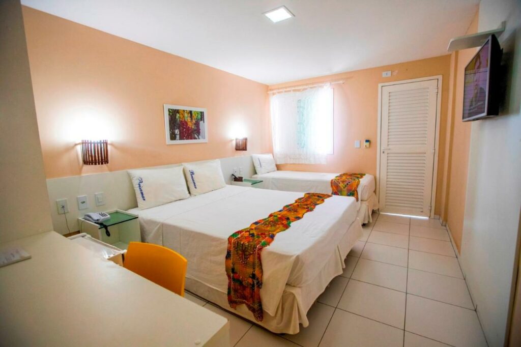 Quarto do Brisamar Hotel Pousada com uma cama de casal e outra de solteiro. Há também uma mesa com cadeiras, janela e uma televisão. Foto para ilustrar post sobre pousadas em Barra de São Miguel.
