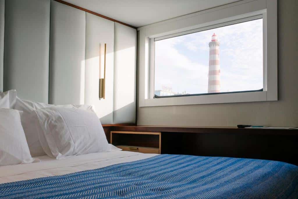 Quarto em barco hotel com cama de casal com colcha azul e branca, bancada marrom ao lado da cama, armário branco atrás cama e janela com vista para um farol. Imagem para ilustrar o post hotéis em Aveiro.